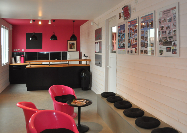 coin salon avec des sections composées d'un grand banc le long d'un mur avec des oreillers noirs ronds, de petits oreillers noirs et des chaises en plastique brillant avec des sièges noirs à côté d'une zone de réception noire et en bois avec un grand mur rose