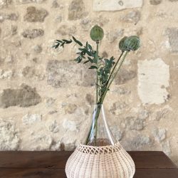 Grand vase fait à la main en verre chiné et rotin avec des plantes vertes, posé sur une table devant un mur en pierre naturelle